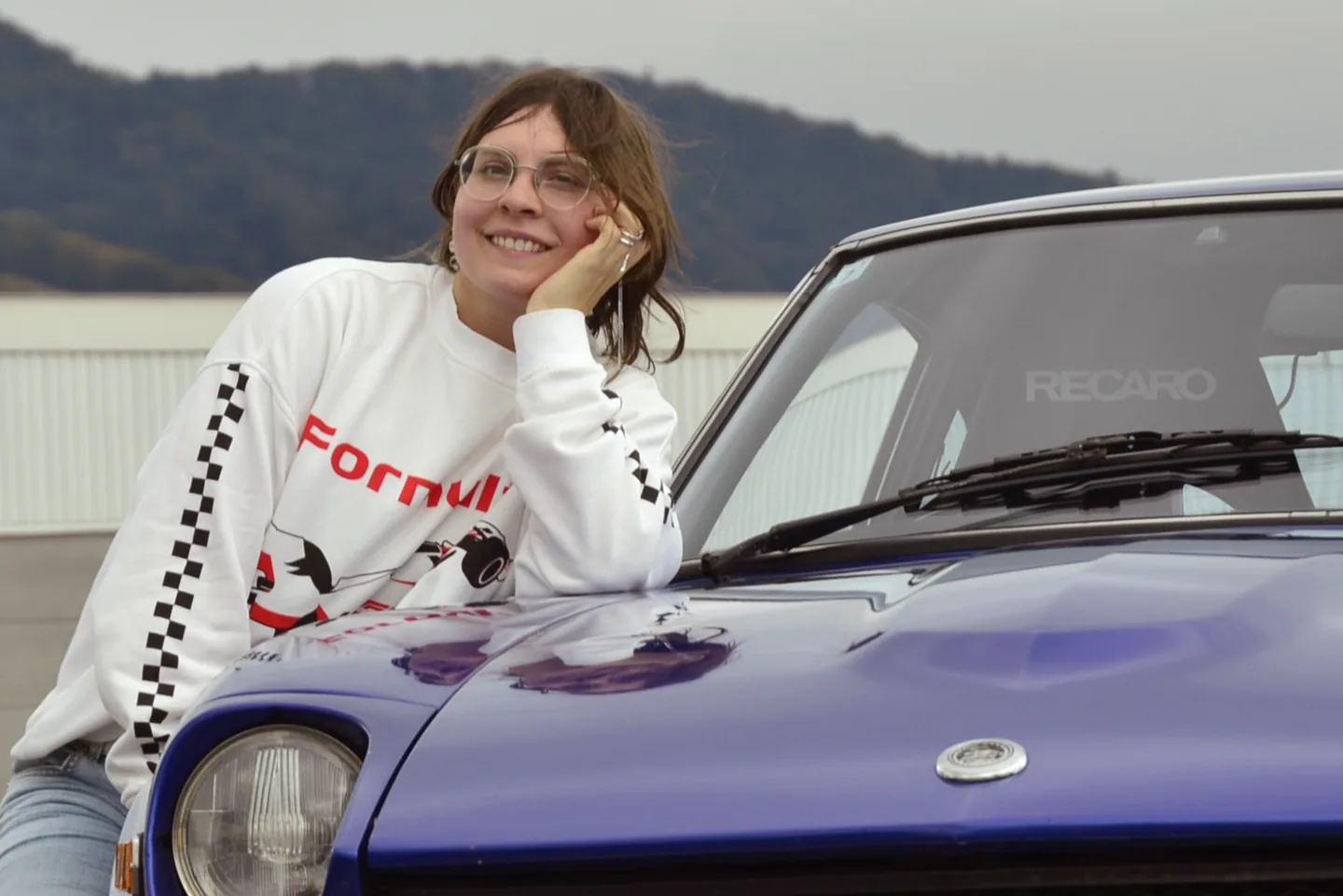 Die Zeichnerin lehnt lächelnd auf der Motorhaube eines blauen Datsun 240Z; im Hintergrund eine bewaldete Hügelkette in Hakone.