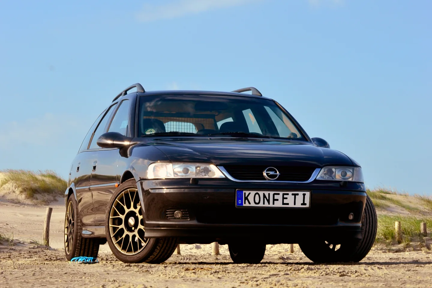 Ein schwarzer Opel Vectra B Caravan mit goldenen Felgen und dem Kennzeichen „KONFETI“ steht in Dreiviertelansicht von vorn auf einem Sandweg zwischen Dünengras.